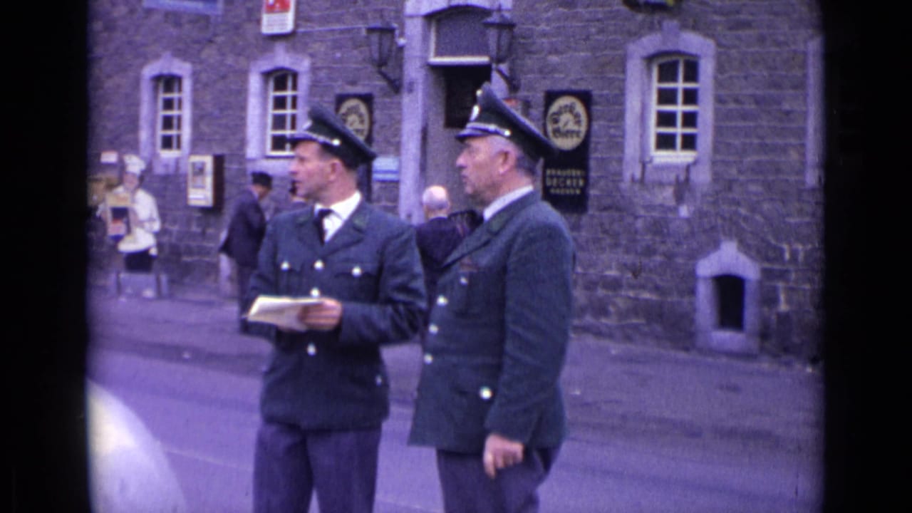 Two uniformed men stand on a street outside a stone building while one holds paper.