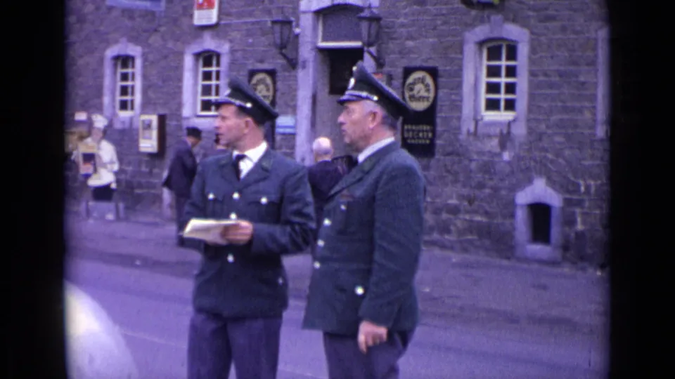 In 1962 Switzerland, two men in dark peaked-cap uniforms stand near a stone building as pedestrians pass behind them.