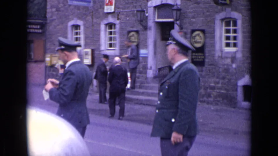 A 1962 Swiss street view shows uniformed men in peaked caps near a stone facade, with pedestrians by the entrance.