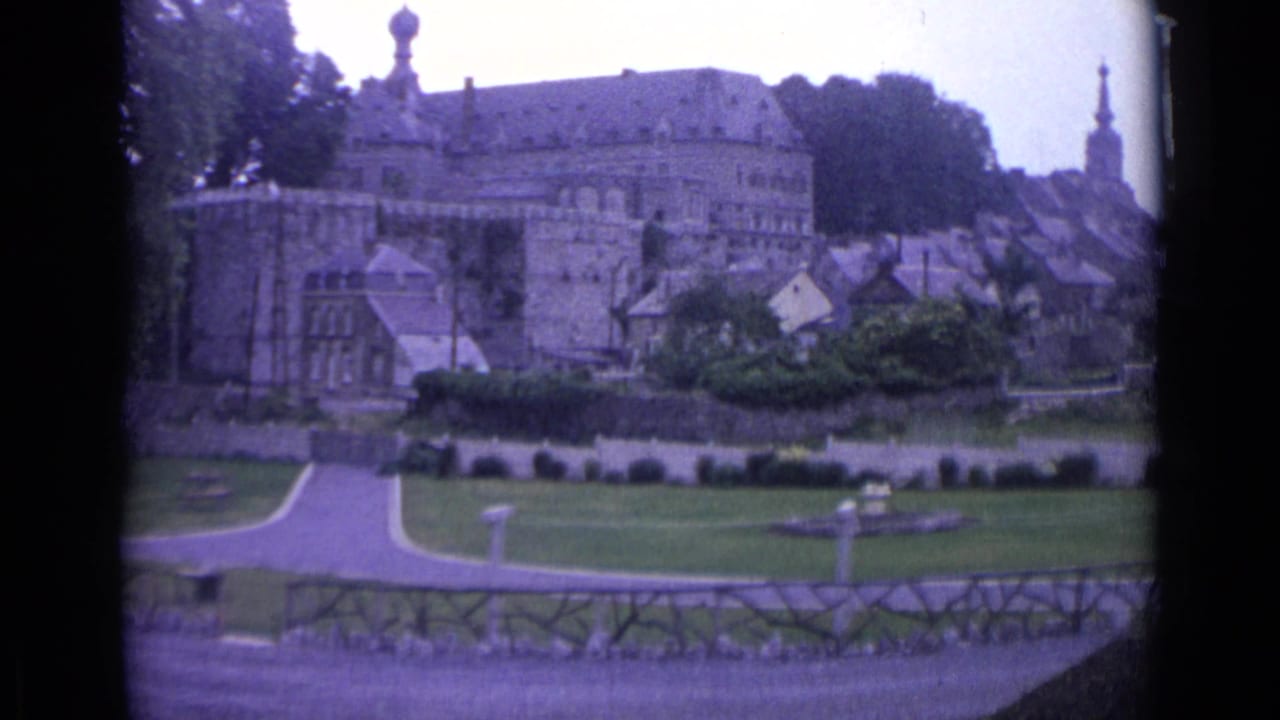 Historic townscape with a large stone building, rooftops, lawns, paths, and a foreground fence.