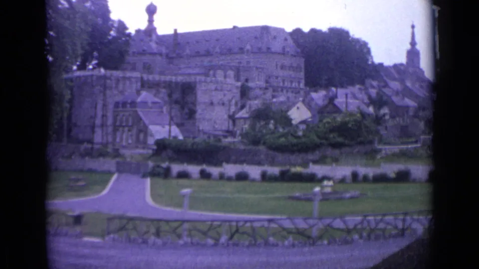 1962 Swiss home-movie frame showing a hilltop stone building, clustered houses, gardens, paths, and a church steeple in soft 8mm color.