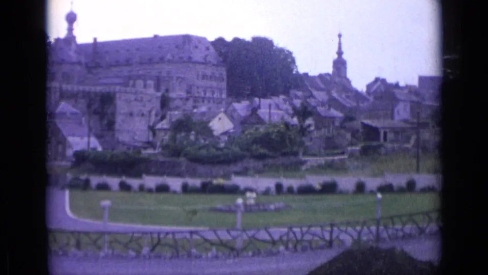 1962 Swiss vacation footage frame with historic buildings above a lawn, a church steeple, garden paths, and a rustic fence in front.