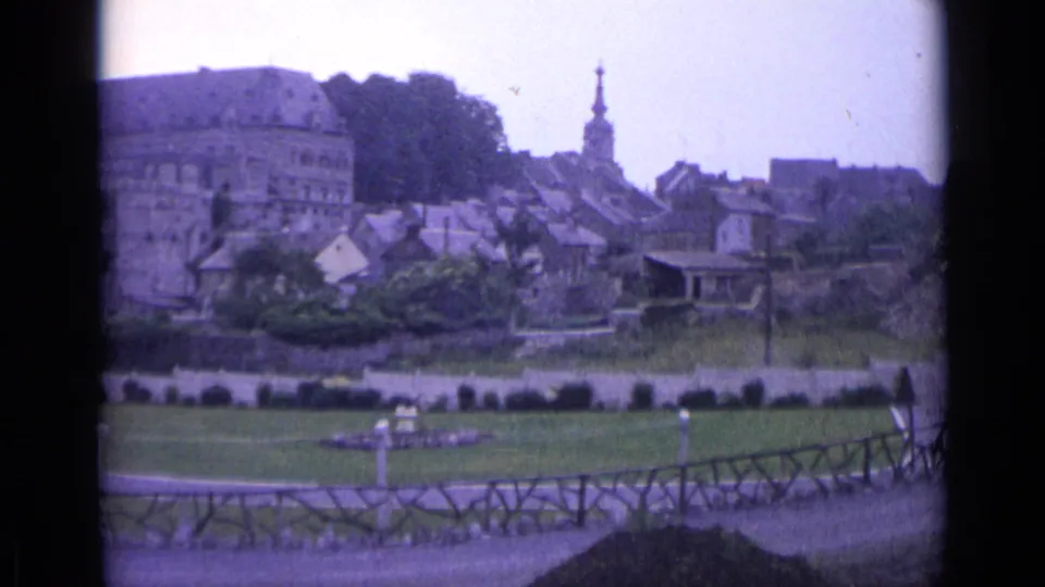 1962 Swiss 8mm frame after a pan, centered on rooftops and a church steeple beyond green lawns, shrubs, and a wooden fence.