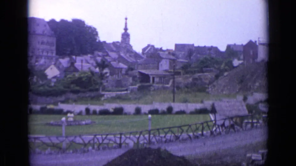 1962 Swiss home-movie frame with rooftops, a church steeple, trees, a lawn, garden beds, and foreground fencing in faded 8mm color.