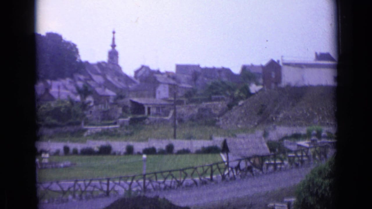 Distant Swiss village view on 8mm film, showing rooftops, a church steeple, grassy foreground, and a fence-lined path.