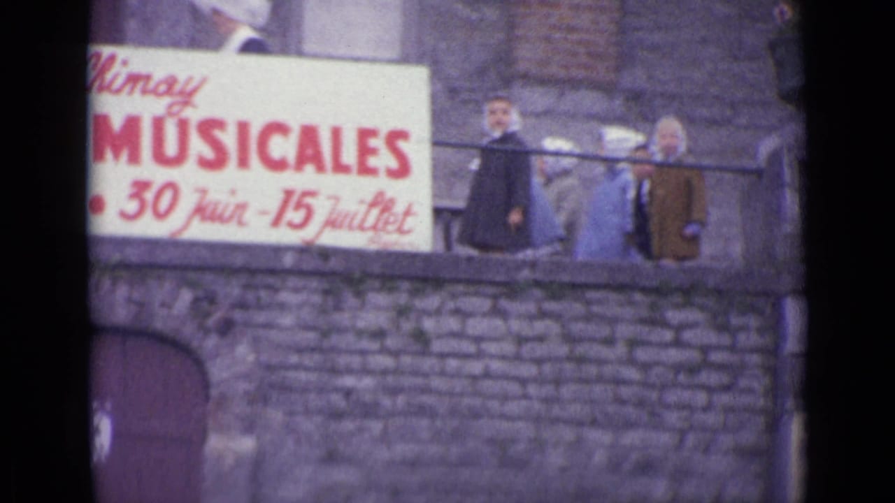8mm frame showing a readable musical festival sign with June and July dates, stone wall, balcony railing, and adults nearby.