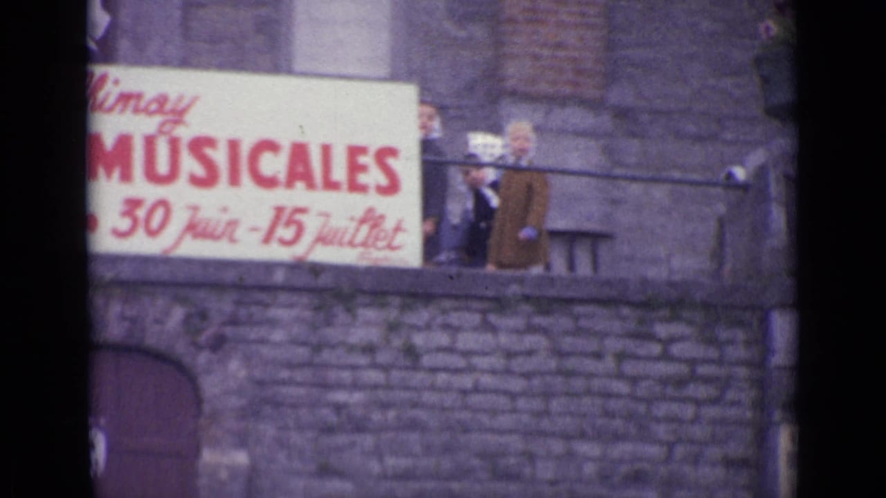 Close 8mm view of the musical festival sign, with adults partly visible behind it and stone architecture around the balcony.