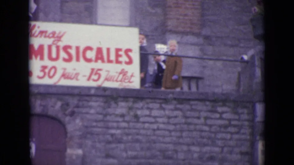 Close 8mm view of the musical festival sign, with adults partly visible behind it and stone architecture around the balcony.