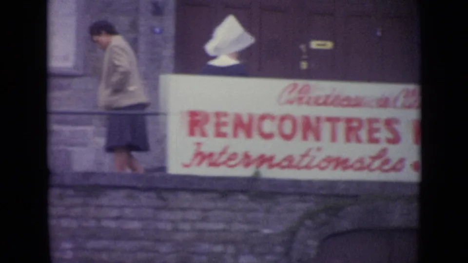 1962 8mm frame cataloged as Switzerland showing an adult passing near the red-lettered musical-event banner on a stone building.