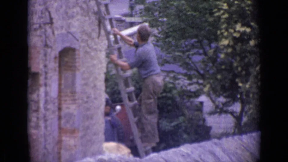 1962 8mm frame cataloged as Switzerland showing an adult working on a ladder near a stone facade and leafy tree.