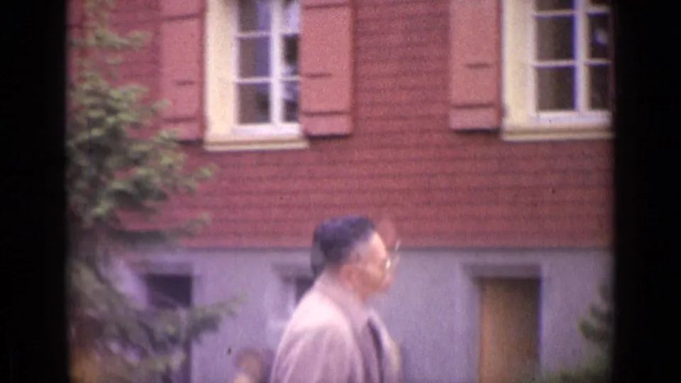 1962 Switzerland frame of an adult man in glasses walking past a red brick building with shutters and foliage.