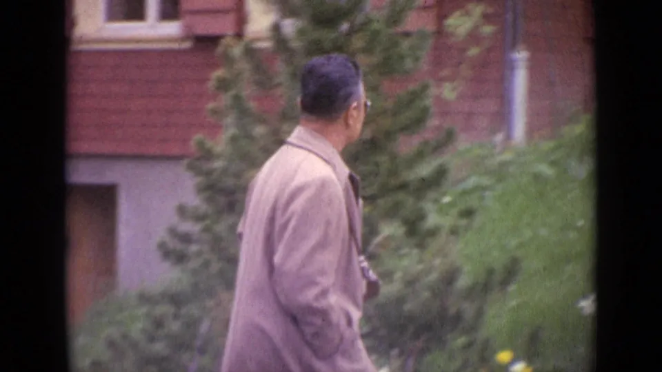 1962 Switzerland frame showing a suited man from behind as he walks beside greenery and a red brick facade.