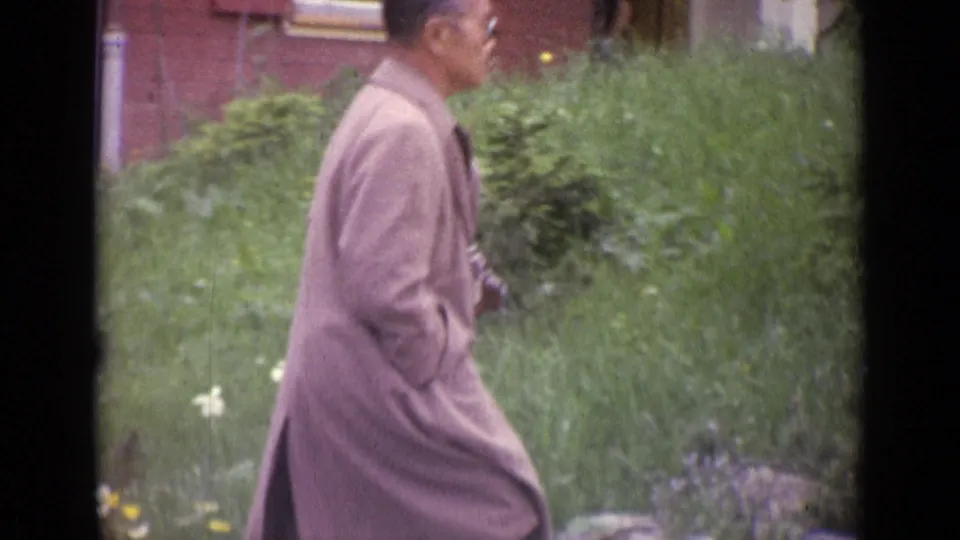 1962 Switzerland frame with a bespectacled man in profile walking past grass, shrubs, and a red brick building.