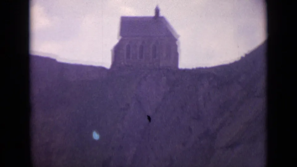 1962 Switzerland 8mm frame showing a small chapel-like hillside building with arched openings under a pale cloudy sky.