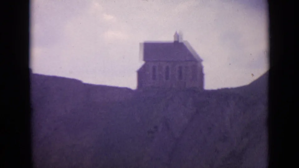 1962 Switzerland 8mm view of a small stone chapel-like building set against a sloping hillside and washed cloudy sky.