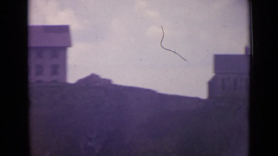 1962 Switzerland 8mm frame during a pan, showing partial hillside buildings separated by a grassy ridge and cloudy sky.
