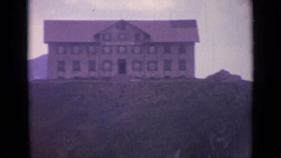 1962 Switzerland 8mm frame showing a larger gabled hillside building with rows of windows and a grassy slope in front.