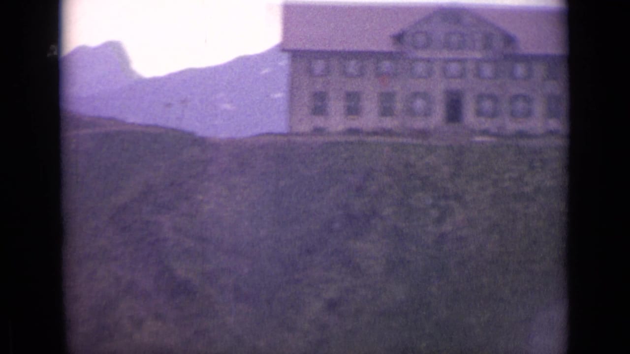 Large alpine building on a grassy hill with mountains behind it.