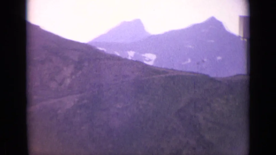Swiss Alps landscape in soft 8mm color, with ridges, distant snow patches, and a pale overexposed sky.