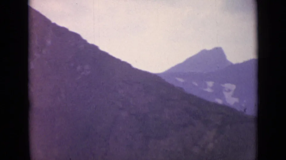 Swiss Alps home-movie frame showing a shadowed slope, distant peak, and snow patches under a pale sky.