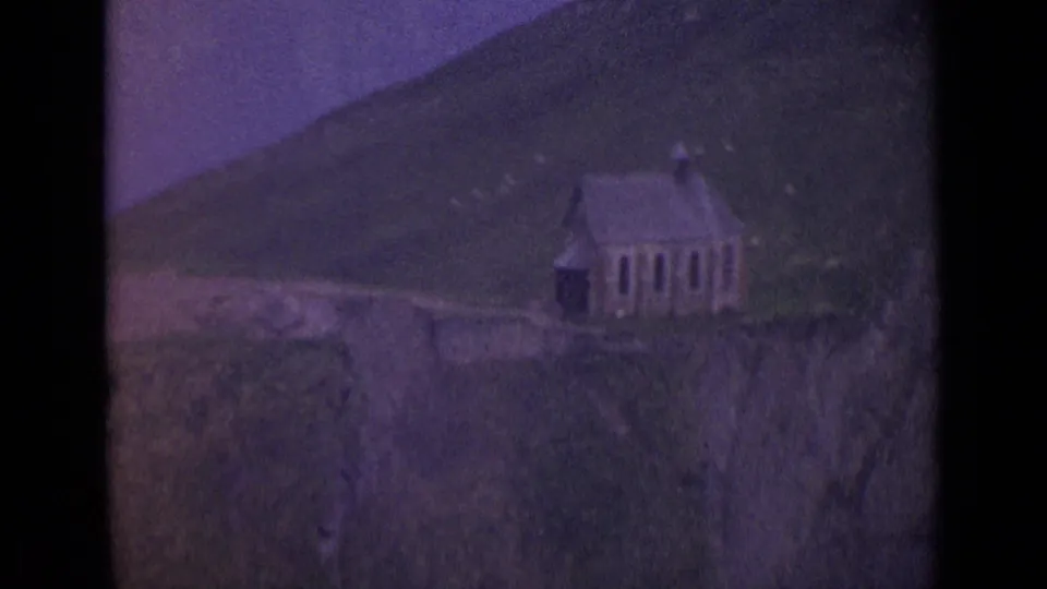 1962 Switzerland 8mm frame showing a small church-like building on a hillside with mountain slopes behind it.