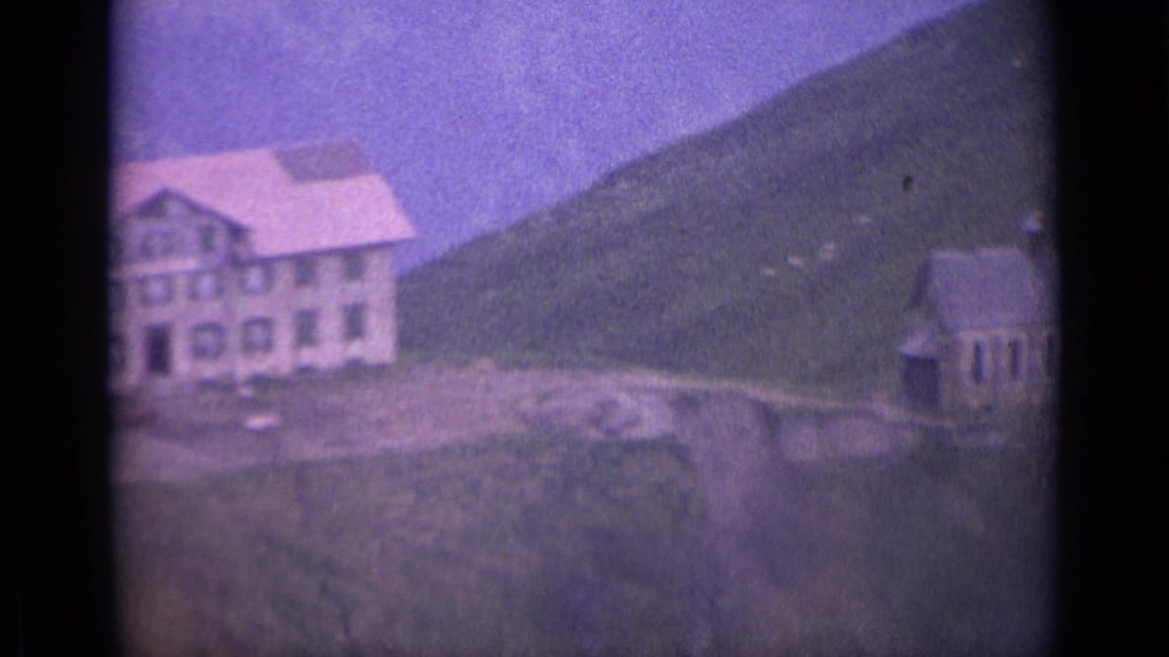 Large pale house and smaller building on a steep green hillside under bright sky.