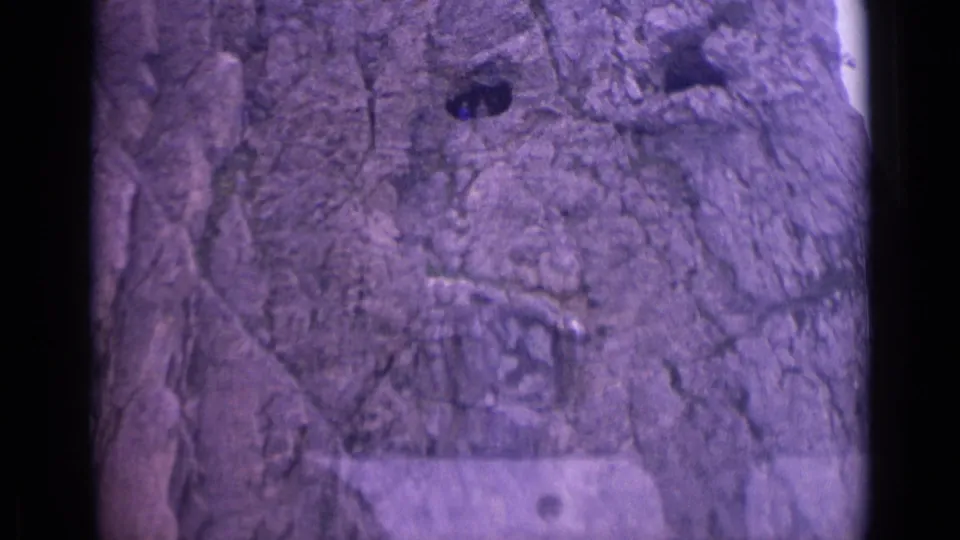 Close 8mm view of a rough Swiss mountain rock face, with dark openings and purple color cast from the film scan.