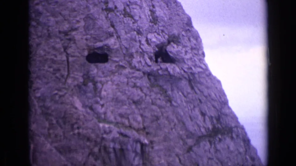 1962 Switzerland 8mm frame showing a craggy alpine rock face with two dark openings and a pale sky beside it.