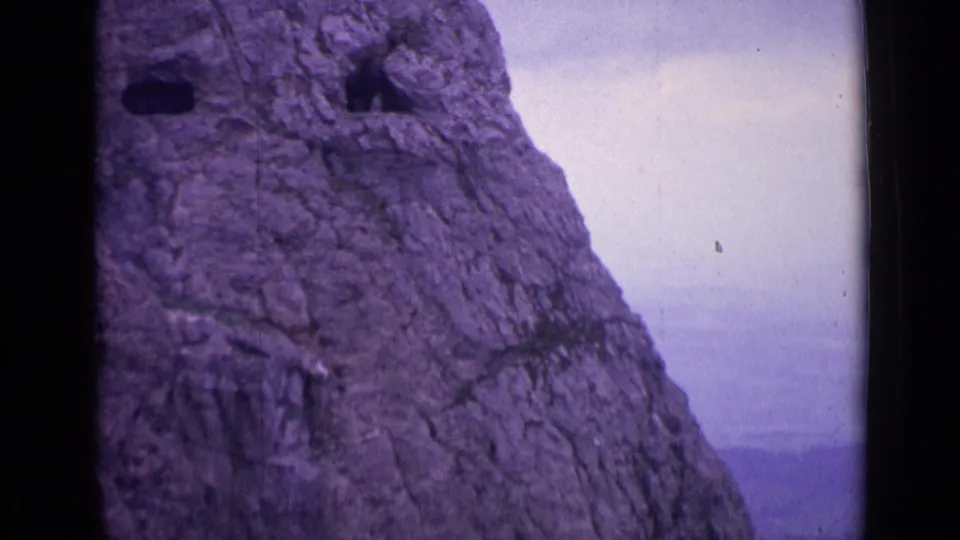 Swiss alpine rock wall on 8mm film, with dark cave-like openings and a hazy pale sky to the right.