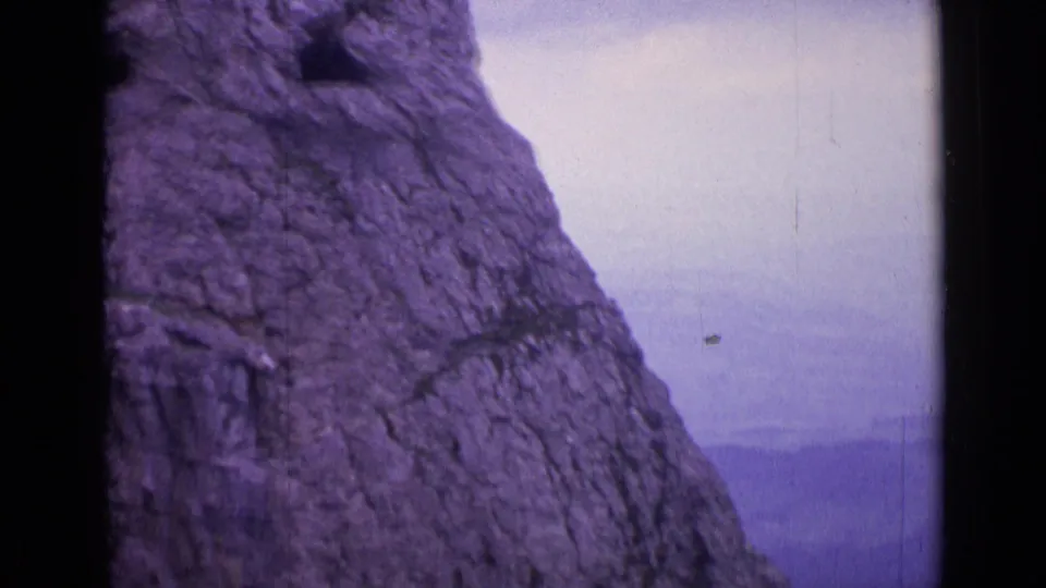 Handheld 1962 Switzerland frame of a steep rock face, a dark opening, and distant haze beyond the cliff.