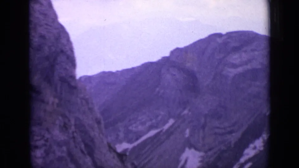 1962 Switzerland home-movie view of a mountain ridge beneath pale sky, framed by dark rocky foreground edges.