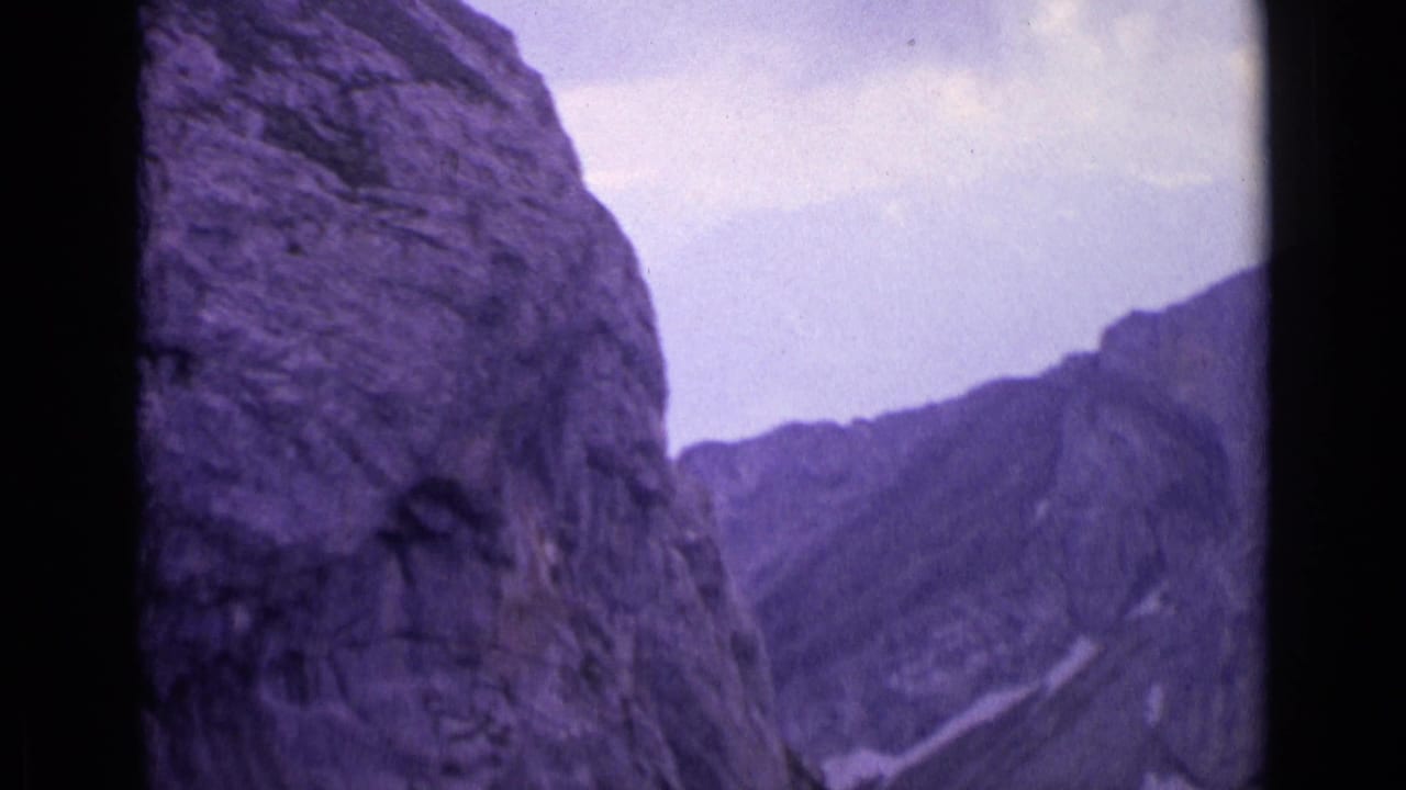 Steep grey rock wall with distant mountain ridge and cloudy sky in a purple-tinted film frame.