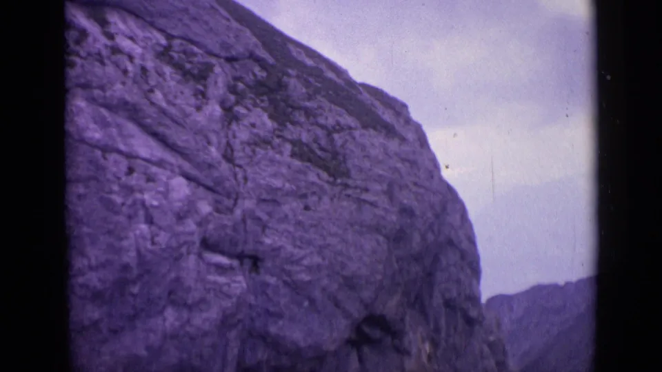1962 Switzerland frame with a massive textured alpine rock formation, cloudy sky, and purple faded 8mm color.