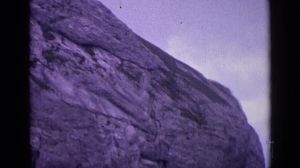 Swiss Alps home-movie frame from 1962 showing a slanted grey rock face beneath cloudy sky with visible 8mm grain.
