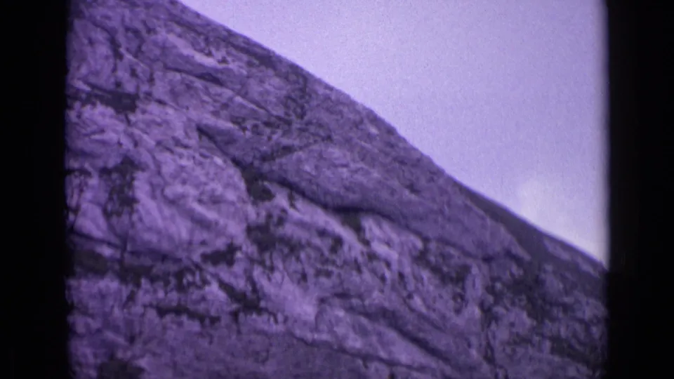 1962 Swiss alpine frame showing a rugged grey rock slope, cloudy sky, and dark 8mm border with purple color shift.