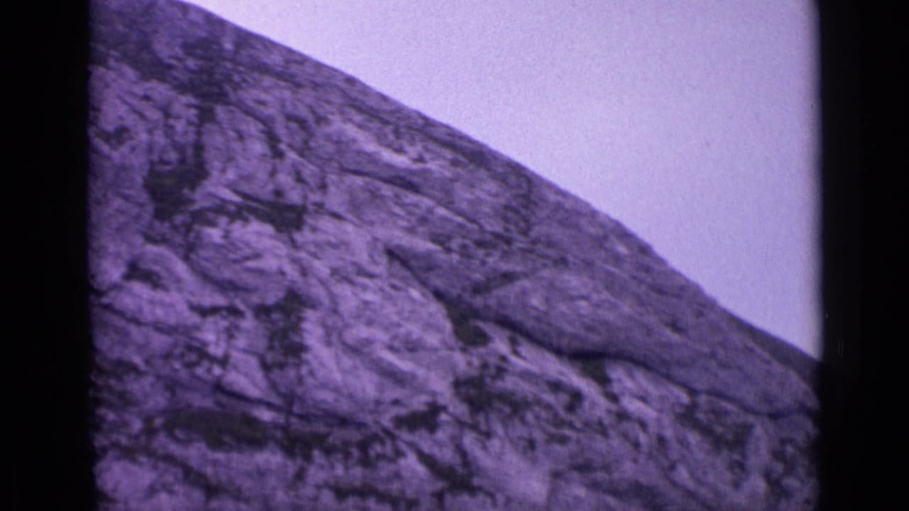 Purple-tinted rocky mountain face under a pale sky with dark film borders.