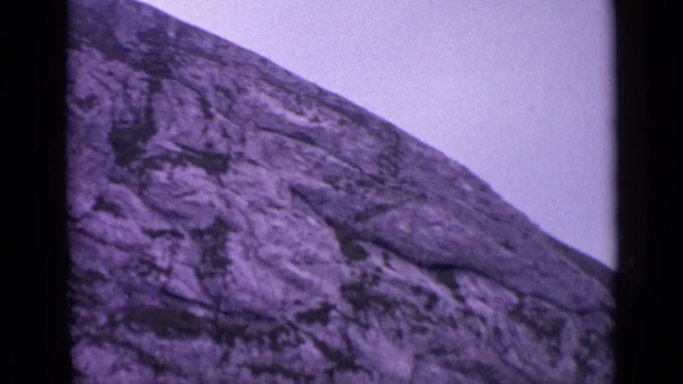 1962 Swiss 8mm frame showing a rugged alpine rock face beneath a pale sky, with visible grain and dark frame edges.
