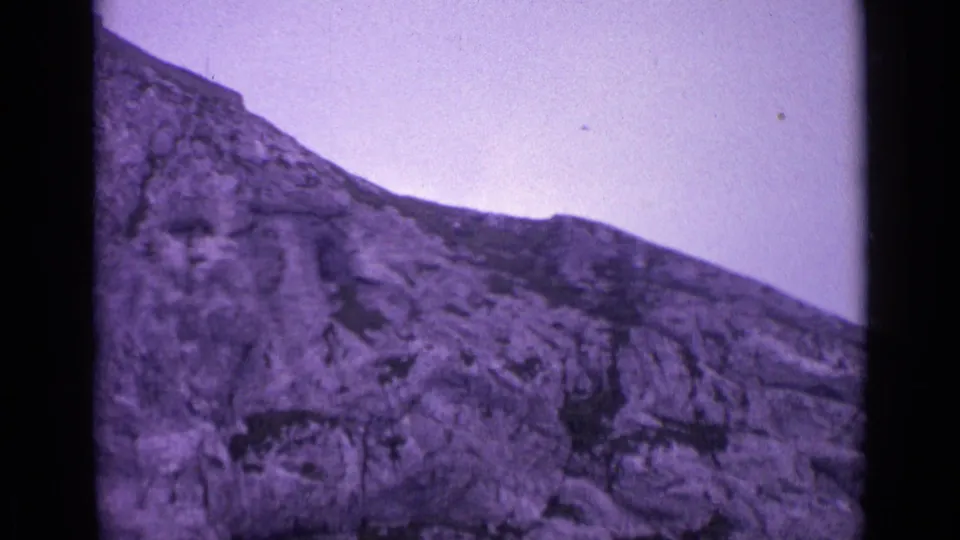 Swiss 8mm home-movie frame showing a steep rocky mountainside, pale sky, and visible mid-century film texture.