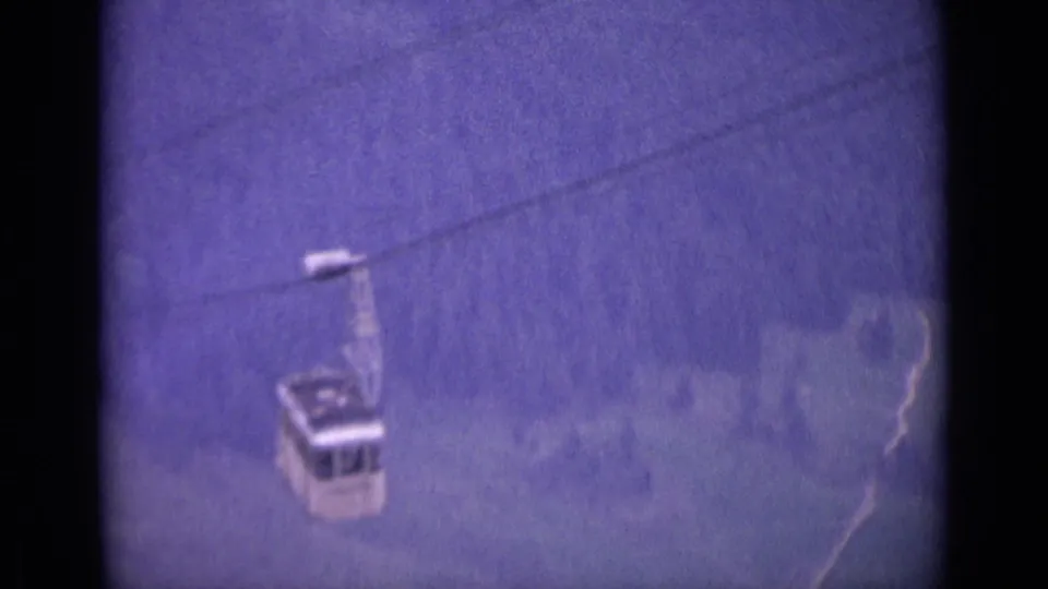 A Swiss cable car appears suspended on a diagonal cable against soft wooded mountain scenery in this 1962 8mm home-movie frame.