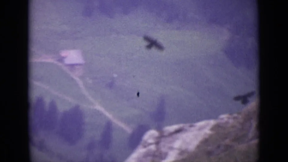 1962 Swiss Alps 8mm frame showing a distant dark bird above a misty mountain valley and rocky foreground.