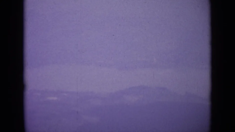 Distant Swiss mountain ridges appear through haze under a purple-tinted sky in the 1962 8mm travel footage.