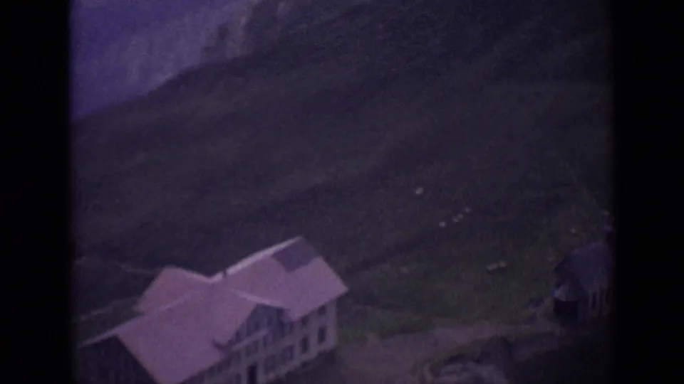 A lodge-like building sits on a green Swiss alpine slope, with mountain ridges and small pale grazing animals nearby.