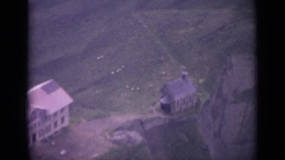 1962 Swiss aerial home-movie frame with a large building, smaller structure, paths, and grazing sheep on alpine terrain.