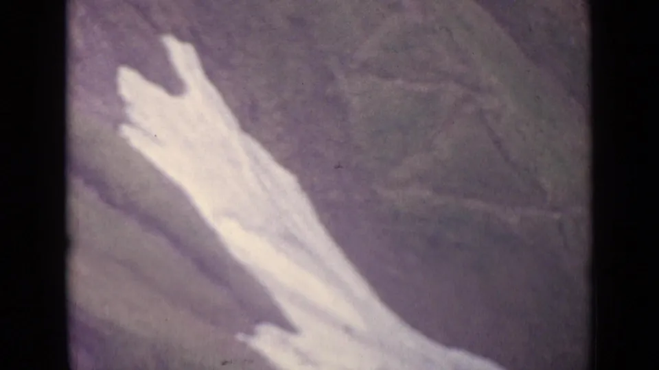 1962 Swiss 8mm frame looking down at a pale snow or rock patch across a grassy alpine hillside.