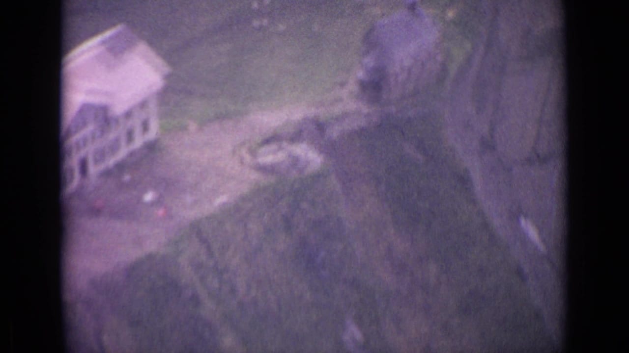 Small buildings sit near a winding path on a grassy mountaintop landscape.