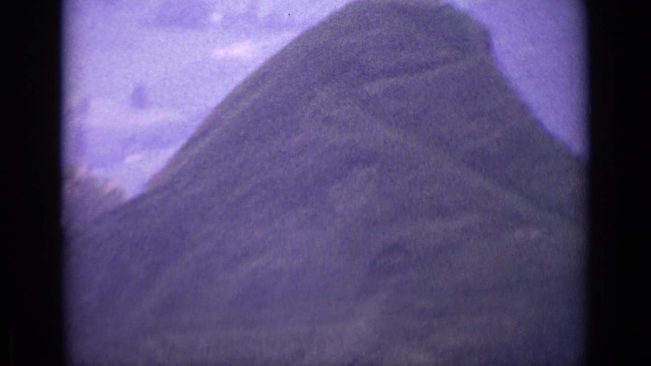 Purple-shifted 1962 Swiss 8mm frame looking over a rounded grassy rise in a mountain landscape.
