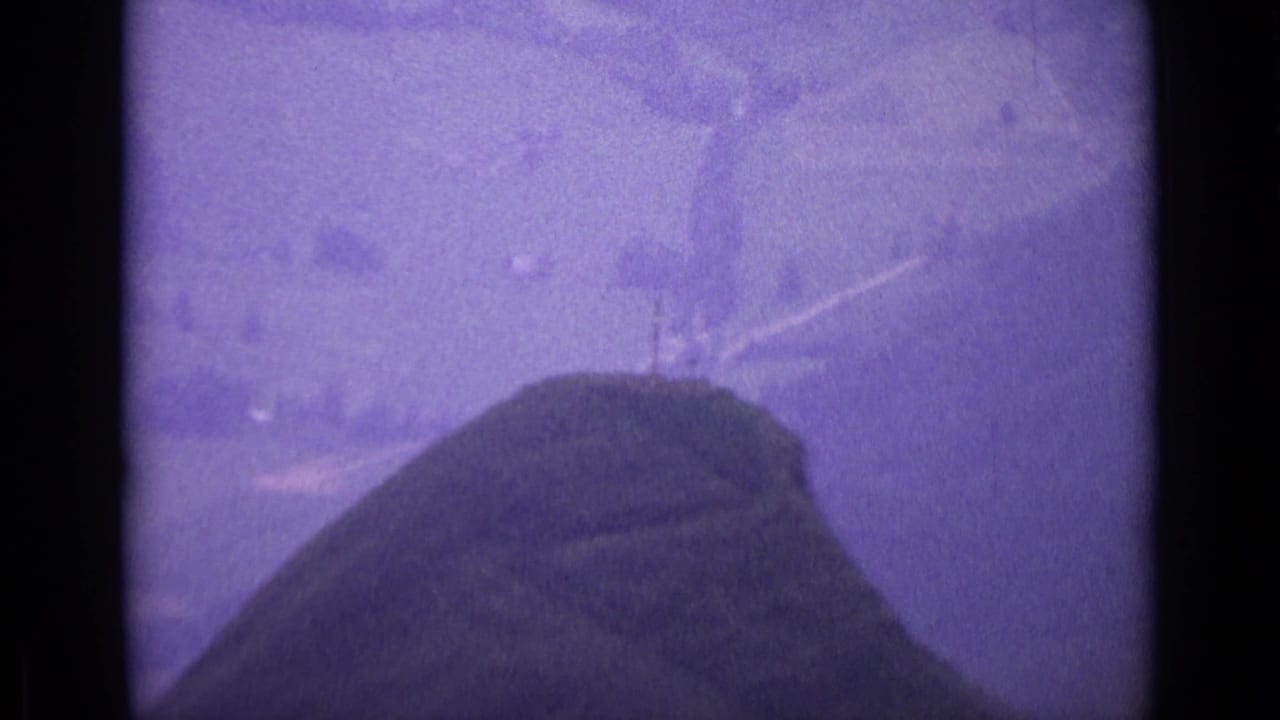 1962 Switzerland home-movie frame showing a rounded mountaintop slope with faint paths and soft distant landscape.