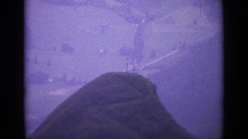 1962 Switzerland home-movie frame showing a rounded mountaintop slope with faint paths and soft distant landscape.