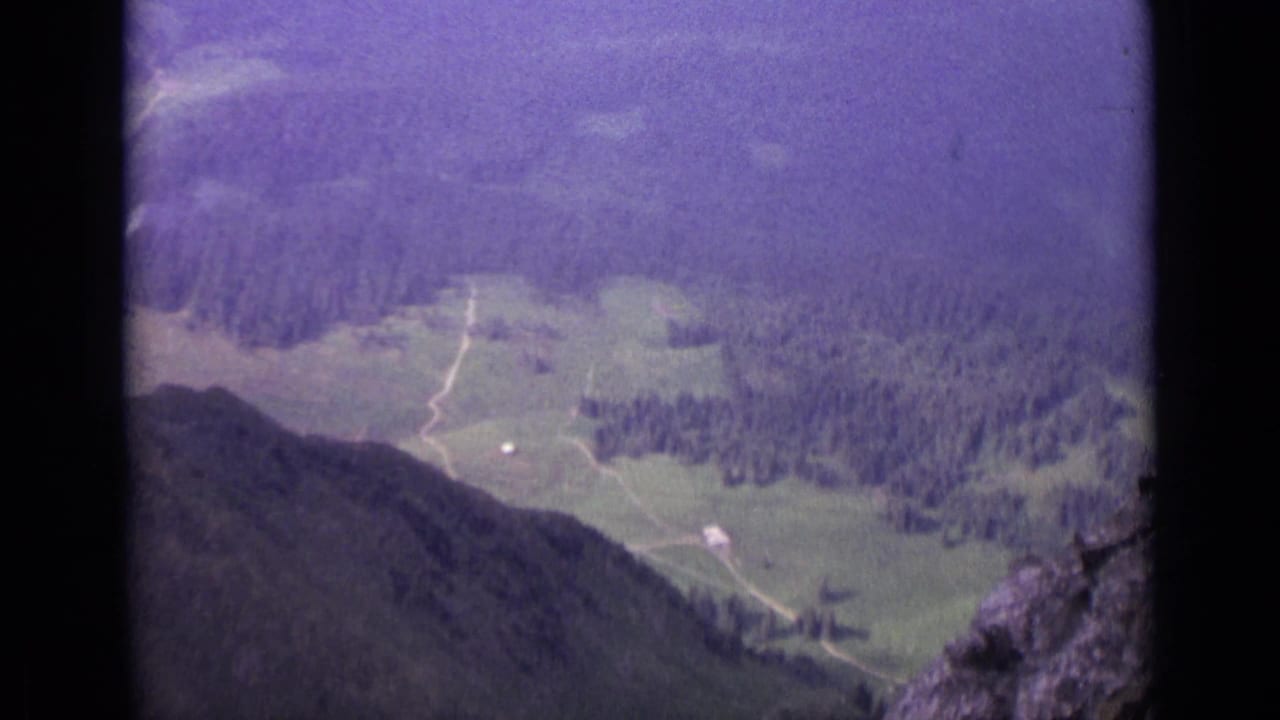 1962 8mm aerial view over a Swiss valley with pasture, dark forest, pale tracks, and steep hillside edges.