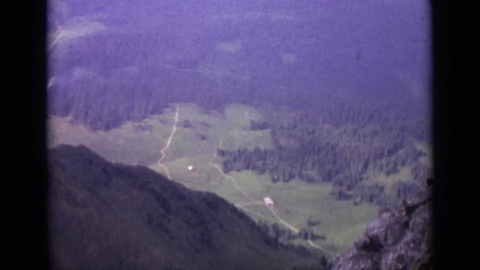 1962 8mm aerial view over a Swiss valley with pasture, dark forest, pale tracks, and steep hillside edges.
